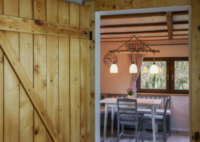 Blick durch eine Holztür in den Essbereich des Ferienhauses Meyer mit einem weißen Esstisch, grauen Stühlen, einer Eckbank und einer charakteristischen Pendelleuchte aus einem Birkenast.