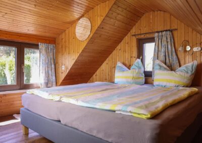 Helles Schlafzimmer unterm Dach mit sichtbarer Holzvertäfelung, einem gemütlichen Doppelbett mit gestreifter Bettwäsche und zwei Fenstern mit Blick ins Grüne im Ferienhaus Meyer.