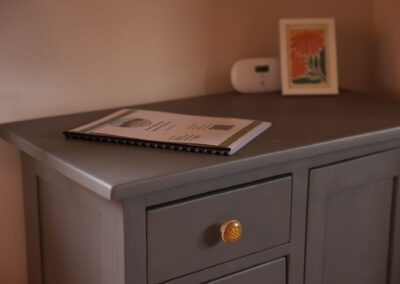 Detailaufnahme einer grauen Kommode mit einem Informationsheft für Gäste, einem verzierten, bunten Keramikknopf an der Schublade im Ferienhaus Meyer.