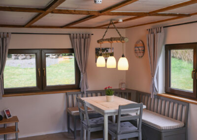 Gemütlicher Essbereich im Ferienhaus Meyer mit einer Eckbank, Stühlen und einem weißen Esstisch, beleuchtet durch eine Pendelleuchte aus einem Baumstamm vor zwei großen Fenstern mit Gartenblick